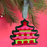 Decorative Christmas chinoiserie ornament shaped like a pagoda hanging on a tree branch with a pink background.