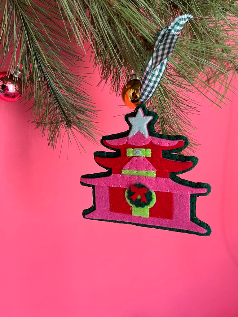 Decorative chinoiserie Christmas ornament shaped like a pagoda with a star on top, hanging from a tree branch against a pink background.