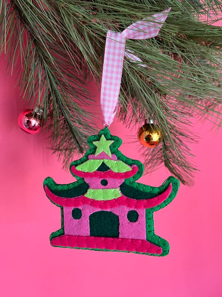 Decorative chinoiserie Christmas ornament shaped like a pagoda with a pink roof and green body, hanging on a tree branch against a pink background.