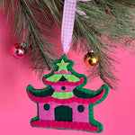 Decorative chinoiserie Christmas ornament shaped like a pagoda with a pink roof and green body, hanging on a tree branch against a pink background.