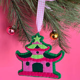 Decorative chinoiserie Christmas ornament shaped like a pagoda with a pink roof and green body, hanging on a tree branch against a pink background.