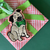 Pug ornament with a green bow tie on a pink and white patterned gift with a green ribbon.
