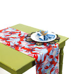 Table runner with a bright red lobster pattern on a blue background styled on a green table with a decorative place setting.