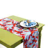 Table runner with a bright red lobster pattern on a blue background styled on a green table with a decorative place setting.