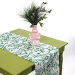 Floral table runner styled on a green table with a pink decorative vase and greenery.