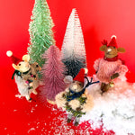 Three festive felt mouse ornaments wearing Santa hats and holiday outfits stand among colorful bottle brush Christmas trees and fake snow, set against a bright red background.