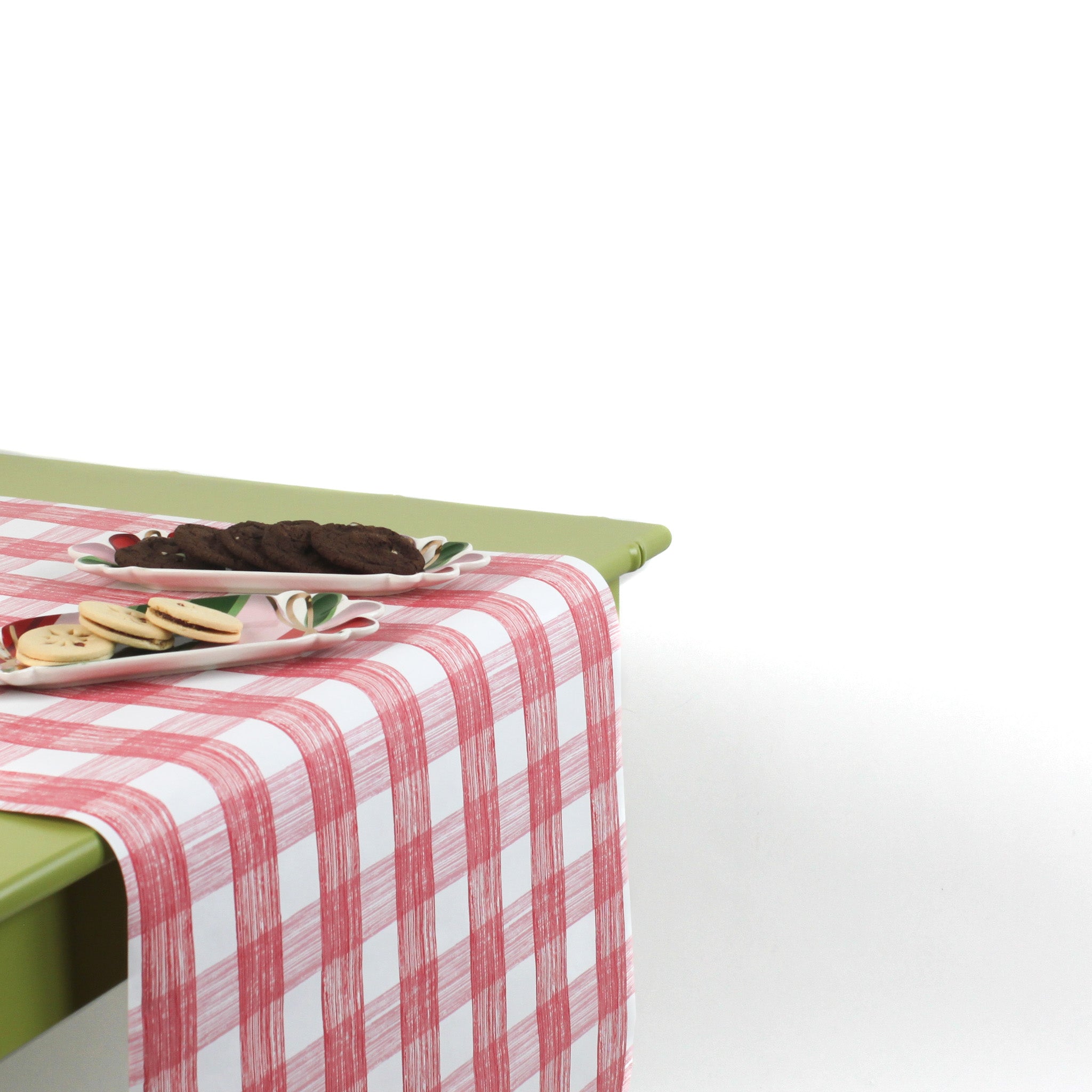 a green table covered with a Gin Lane Pink Gingham Paper Table Runner with a ceramic plate and cookies on top of it