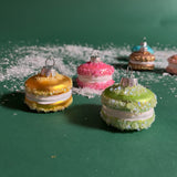 four glass macaron ornaments in green,yellow,and pink and gainst a green snowy background.