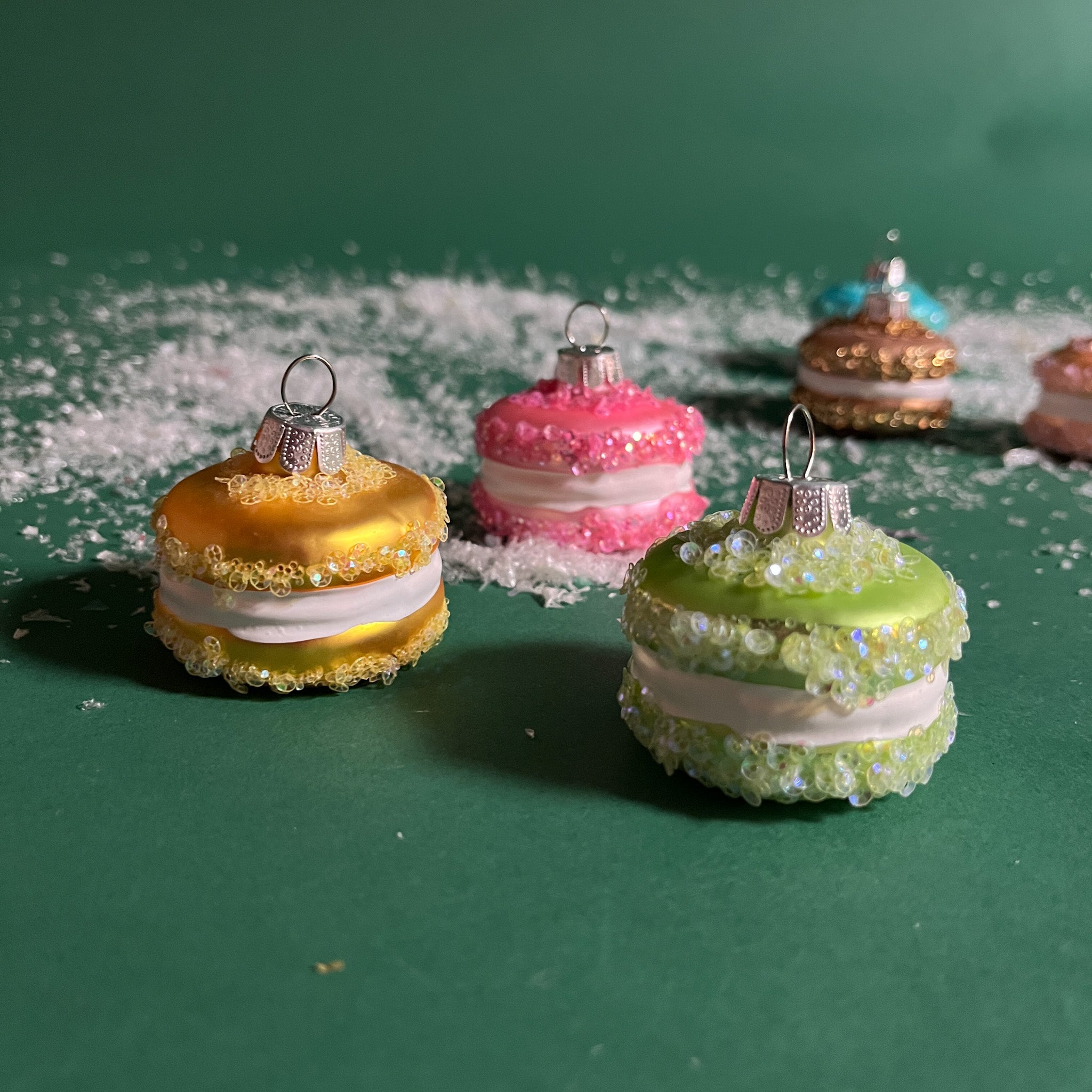 four glass macaron ornaments in green,yellow,and pink and gainst a green snowy background.