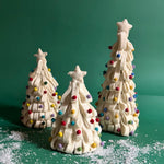 a set of three, white Looped Felt Christmas Tree Table Decorations with colourful bauble decorations, topped with stars against a green, snow scattered background. 