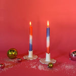lighted 2 colorful taper candles in a mix of white, blue and orange in candle holders and with red background