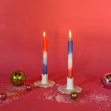 lighted 2 colorful taper candles in a mix of white, blue and orange in candle holders and with red background