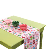 Pink table runner with colorful Christmas ornament patterns displayed on a green table with decorative bowls.