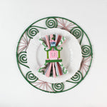 Lifestyle shot of the Shell rattan placemat/charger in pink and green with a white plate resting on it, holding a napkin with pagoda napkin ring.