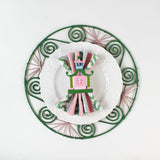 Lifestyle shot of the Shell rattan placemat/charger in pink and green with a white plate resting on it, holding a napkin with pagoda napkin ring.
