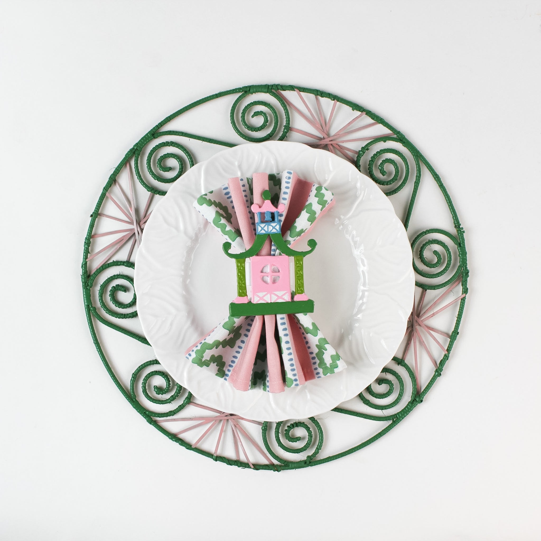 Lifestyle shot of the Shell rattan placemat/charger in pink and green with a white plate resting on it, holding a napkin with pagoda napkin ring.