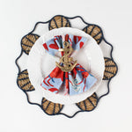 Lifestyle shot of the Shell rattan placemat/charger in navy blue with a white plate resting on it, holding a gingham napkin with gold anchor napkin ring.