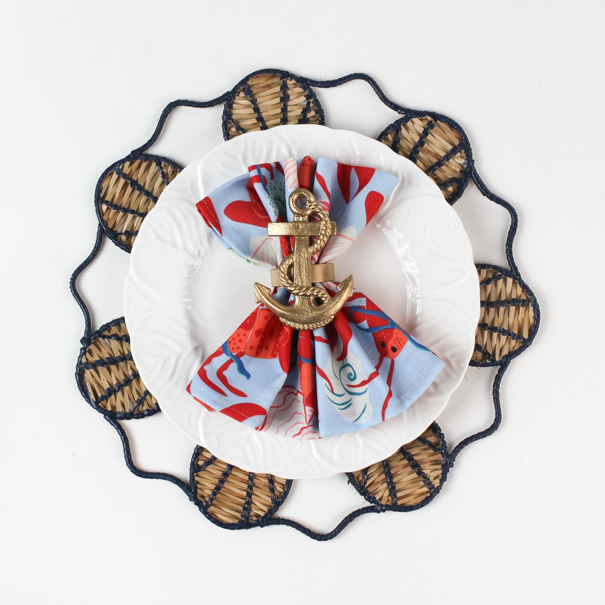 Lifestyle shot of the Shell rattan placemat/charger in navy blue with a white plate resting on it, holding a gingham napkin with gold anchor napkin ring.