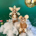 Two felt mouse ornaments dressed as snowflakes stand on fake snow beneath a gold disco ball, surrounded by scattered candy on a green background.
