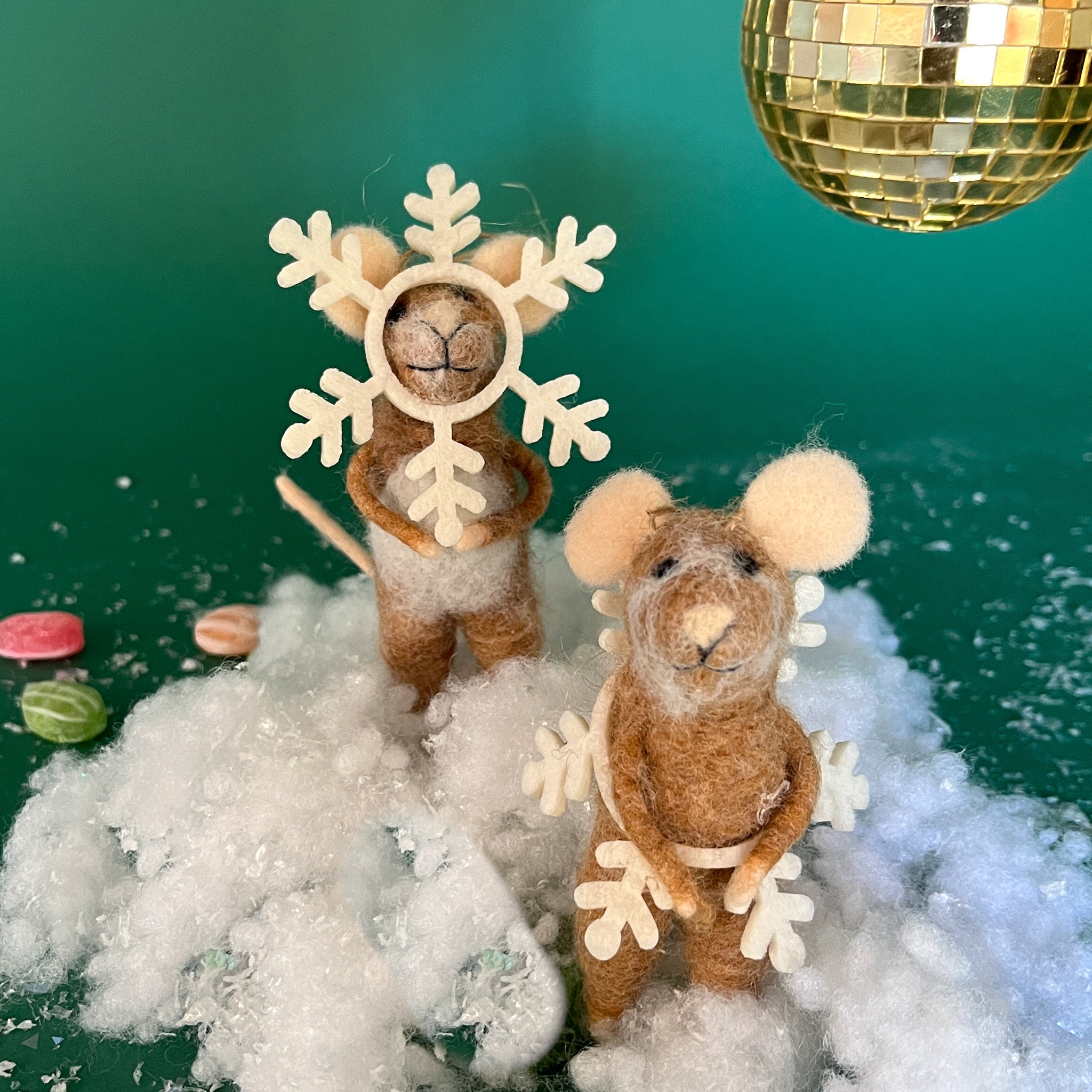 Two felt mouse ornaments dressed as snowflakes stand on fake snow beneath a gold disco ball, surrounded by scattered candy on a green background.
