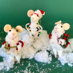 Three white felt mouse ornaments dressed in cozy winter outfits, one wrapped in colorful holiday lights, one holding a Christmas wreath, and one holding a festive ornament while standing on a fake snow