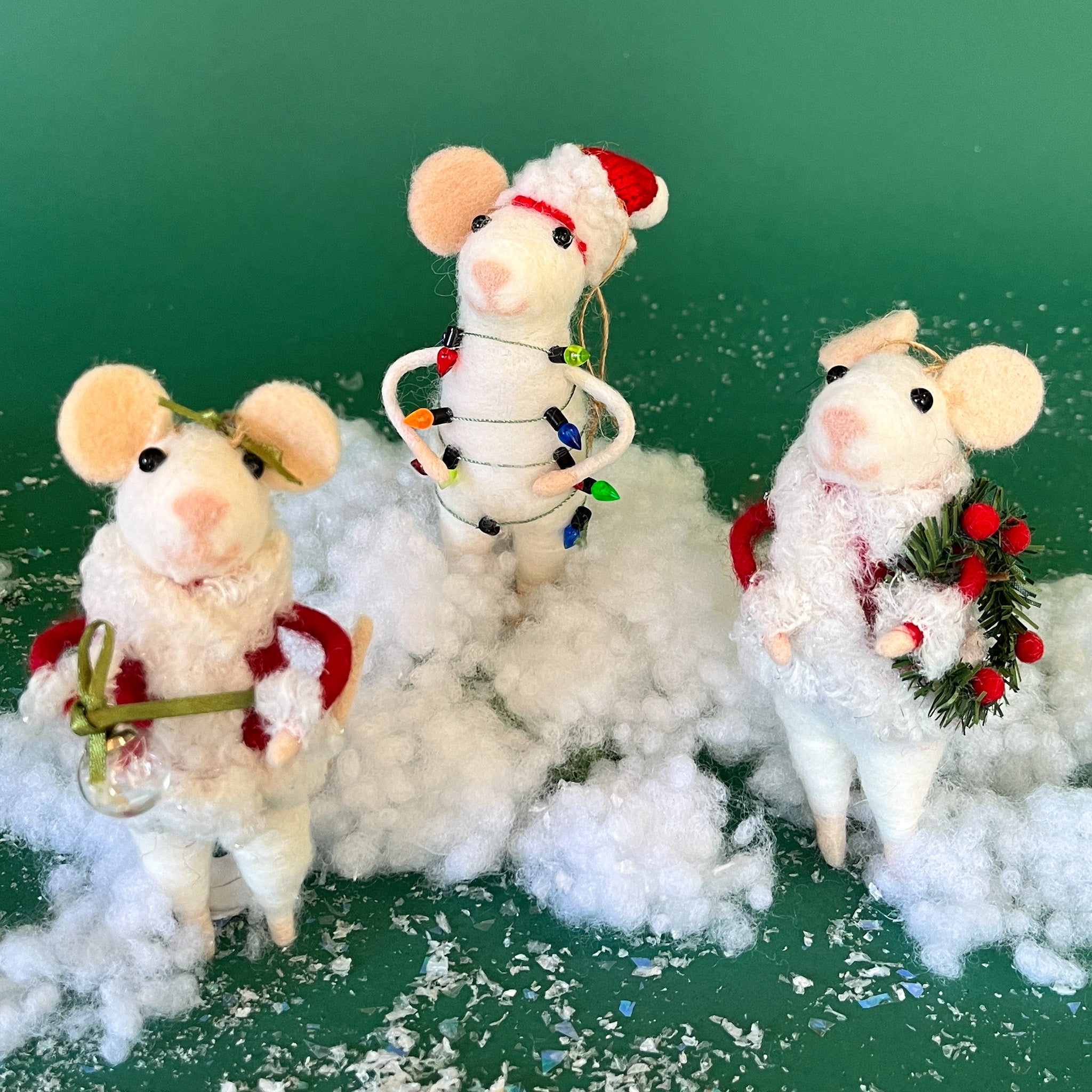 Three white felt mouse ornaments dressed in cozy winter outfits, one wrapped in colorful holiday lights, one holding a Christmas wreath, and one holding a festive ornament while standing on a fake snow
