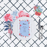 A styled floral table setting featuring a die cut floral paper placemat, white plate, floral menu card, decorative place card, and silver utensils on a navy trellis patterned background.