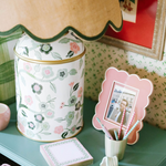 a pink and green chinoiserie lamp with a green trimmed scalloped rattan shade sitting on a blue nightstand. 