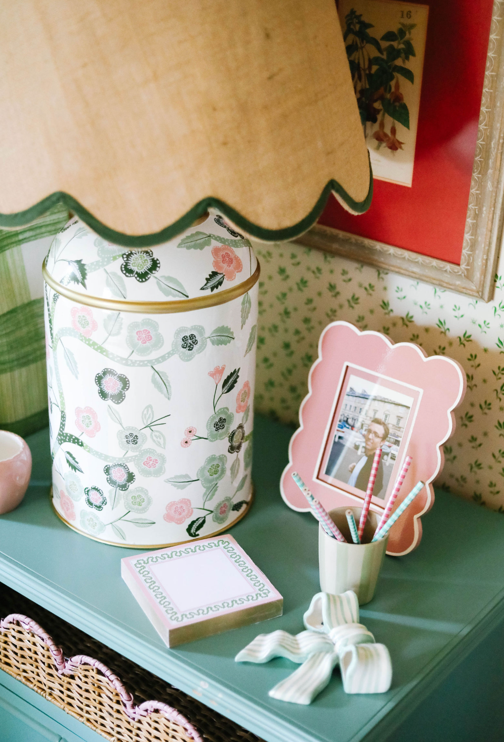 a pink and green chinoiserie lamp with a green trimmed scalloped rattan shade sitting on a blue nightstand. 