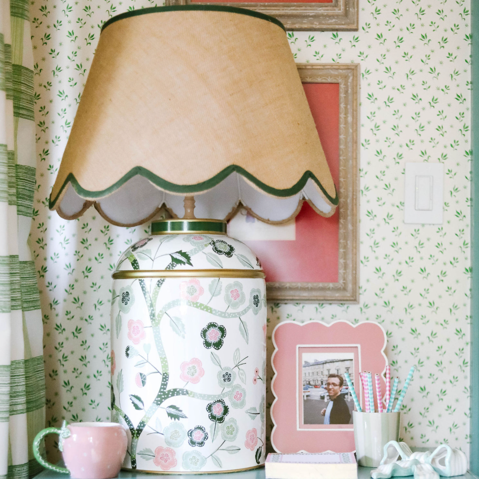 a pink and green chinoiserie lamp with a green trimmed scalloped rattan shade sitting on a blue nightstand. 