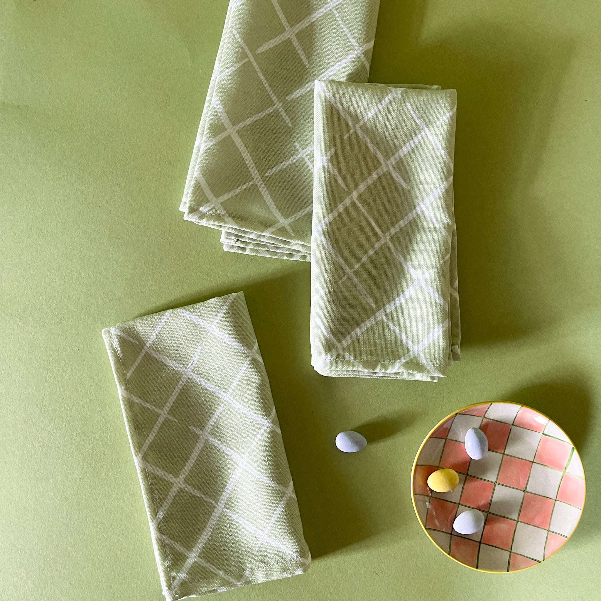 three light green dinner napkins in bahama court trellis print madcap cottage fabric next to a gingham plate with small candy eggs on it