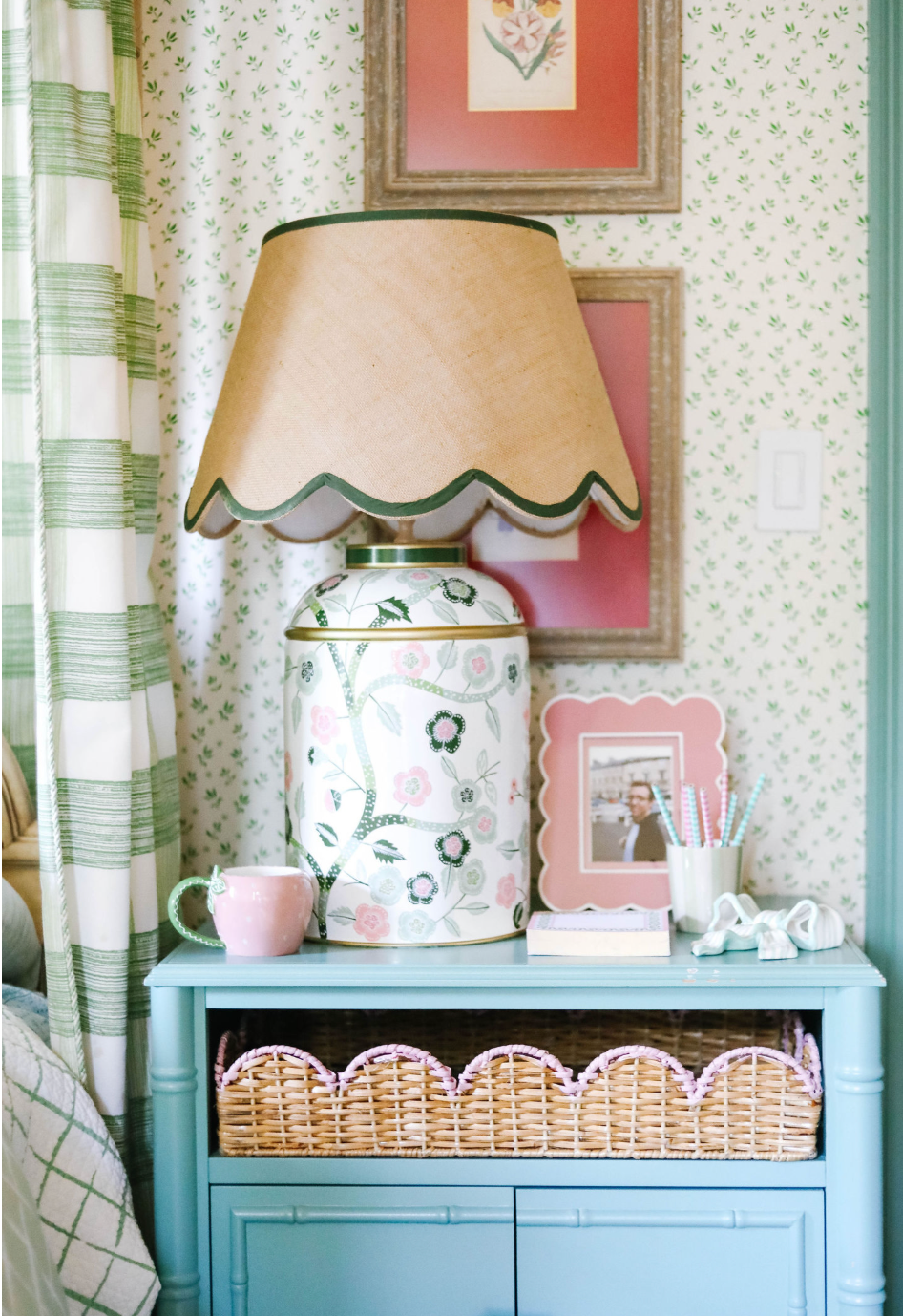 a pink and green chinoiserie lamp with a green trimmed scalloped rattan shade sitting on a blue nightstand. 