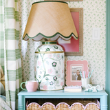 a pink and green chinoiserie lamp with a green trimmed scalloped rattan shade sitting on a blue nightstand. 