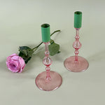 two pink glass candle holders with green tops with a pink rose behind them