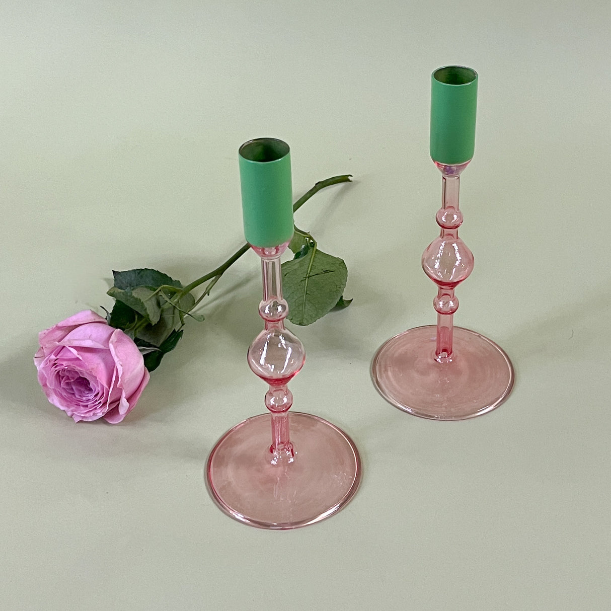 two pink glass candle holders with green tops with a pink rose behind them