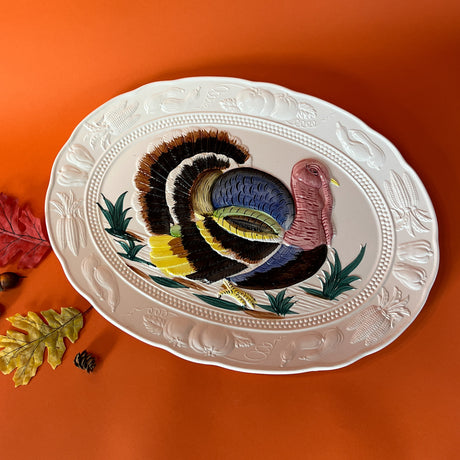 An oversized oval ceramic vintage turkey platter with a vibrantly colored, three-dimensional turkey design in the center, surrounded by a leafy pattern on the rim. The platter is displayed on an orange background with a few autumn leaves scattered around it.