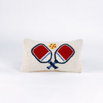 Rectangular white pillow with a hooked wool design of two crossed red and blue pickleball paddles and a yellow pickleball above them.