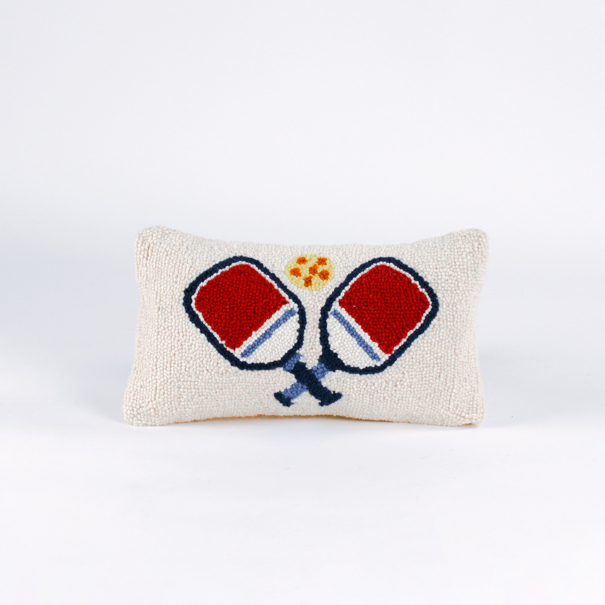 Rectangular white pillow with a hooked wool design of two crossed red and blue pickleball paddles and a yellow pickleball above them.