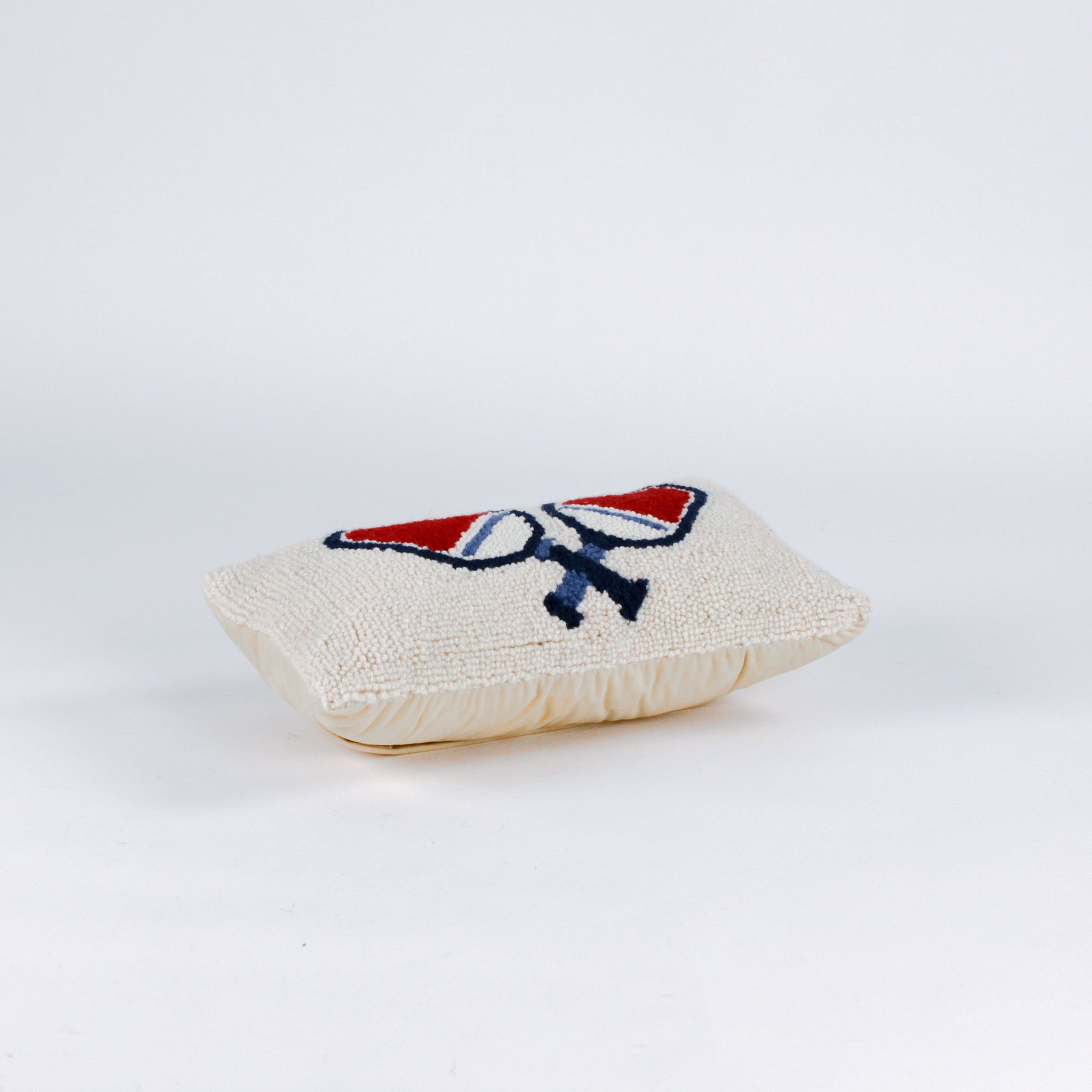 side of a Rectangular white pillow with a hooked wool design of two crossed red and blue pickleball paddles and a yellow pickleball above them.