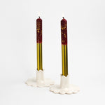 Two white scalloped ceramic candle holders holding tall red and gold taper candles with lit flames, photographed on a white background.