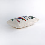side of a Rectangular white pillow with a hooked wool design featuring colorful skis, poles, boots, and scarves hanging on a wooden rack, evoking a cozy winter ski lodge feel.