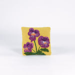 Square decorative pillow with a yellow background featuring a hooked wool design of three purple pansy flowers with green stems and leaves.