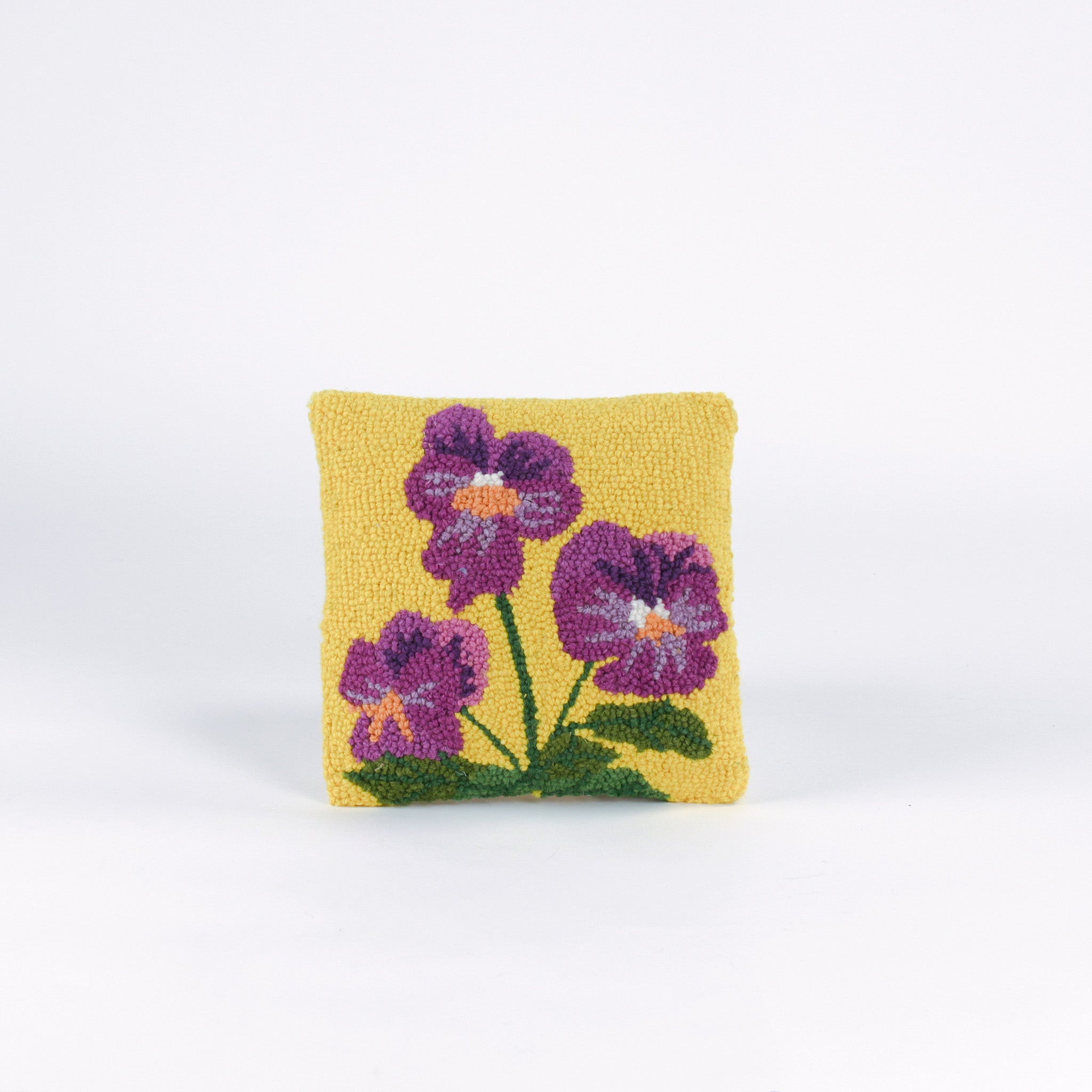 Square decorative pillow with a yellow background featuring a hooked wool design of three purple pansy flowers with green stems and leaves.