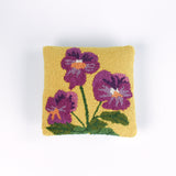 Square decorative pillow with a yellow background featuring a hooked wool design of three purple pansy flowers with green stems and leaves.