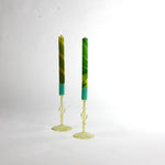 Pair of yellow glass taper candle holders with turned stems and round bases, holding tall green taper candles with lit flames, photographed on a white background.