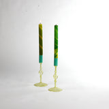 Pair of yellow glass taper candle holders with turned stems and round bases, holding tall green taper candles with lit flames, photographed on a white background.