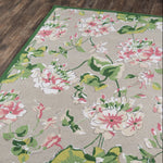 A corner detail of a pink and green rug in a white and pink floral pattern on a grey background and green border.
