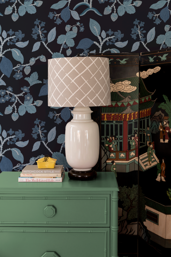 a beige lamp on a green chest of drawers against a blue patterned wallpaper wall.
