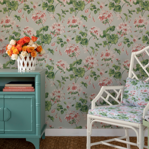 A room with a light blue side table, white chair and geranium wallpaper featuring white and pink geraniums on a light blue background. 