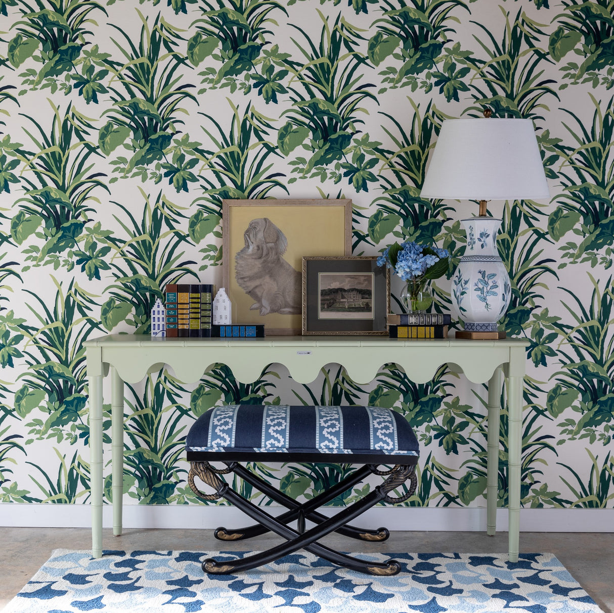 a light green console table topped with a lamp and decorative objects against a background of palm leaf print wallpaper